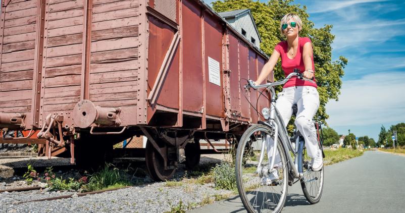 Femme roulant en vélo sur la ligne L156 du RAVel et passant près d'un ancien wagon de train